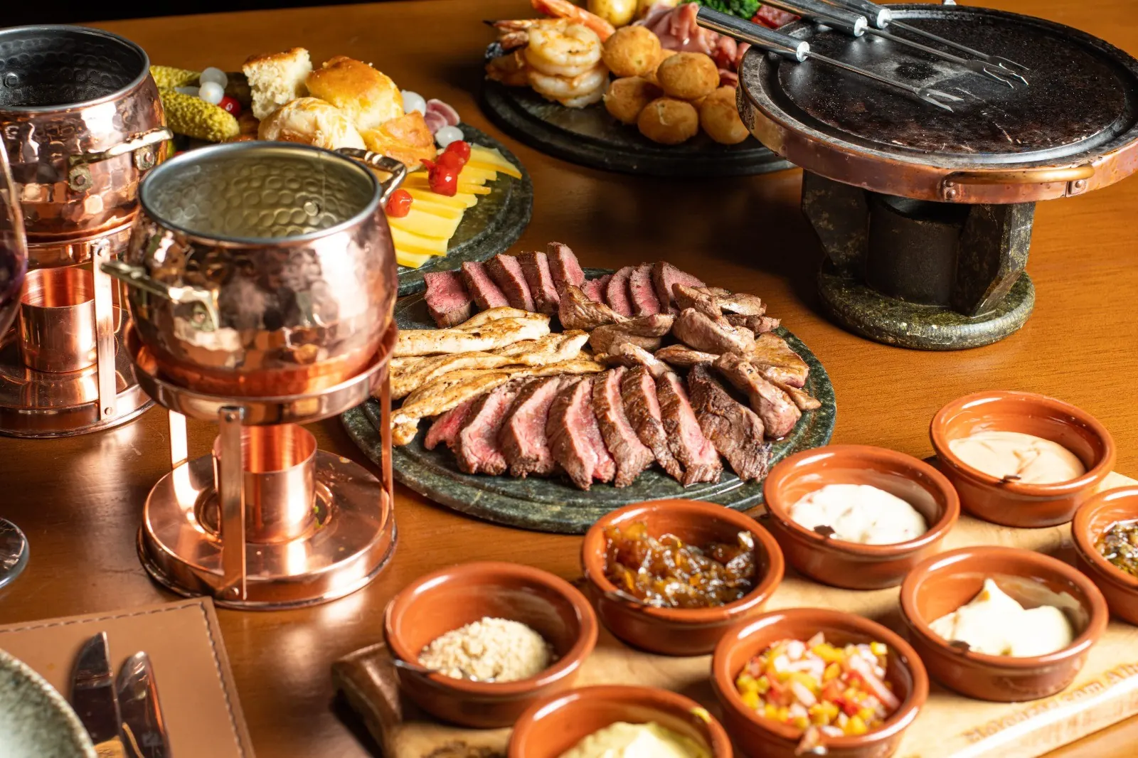 Panelas de cobre com carnes fatiadas e molhos na mesa do jantar